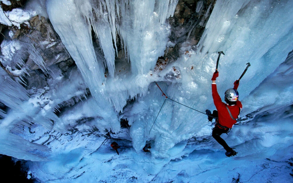 Climber in red jacket scaling frozen waterfall with blue and white icicles winter adventure wallpaper