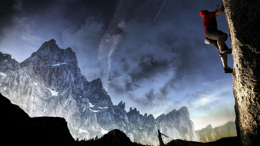 Man climbing rugged rock face with icy mountain backdrop under moody cloudy sky wallpaper