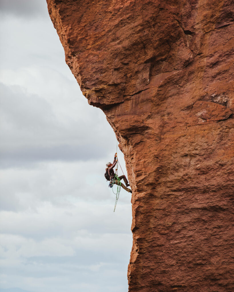 Climber ascending towering red rock cliff under dramatic cloudy sky adventure outdoors wallpaper