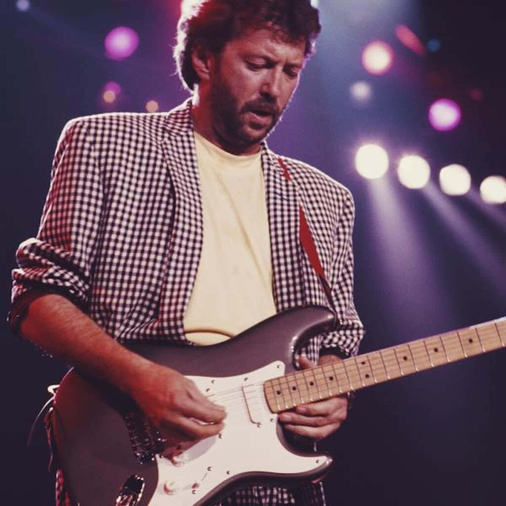 Eric Clapton playing guitar passionately on stage with vibrant concert lights background Wallpaper