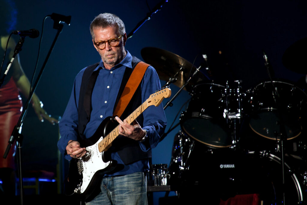 Eric Clapton passionately playing electric guitar on stage with concert lighting and drums background Wallpaper