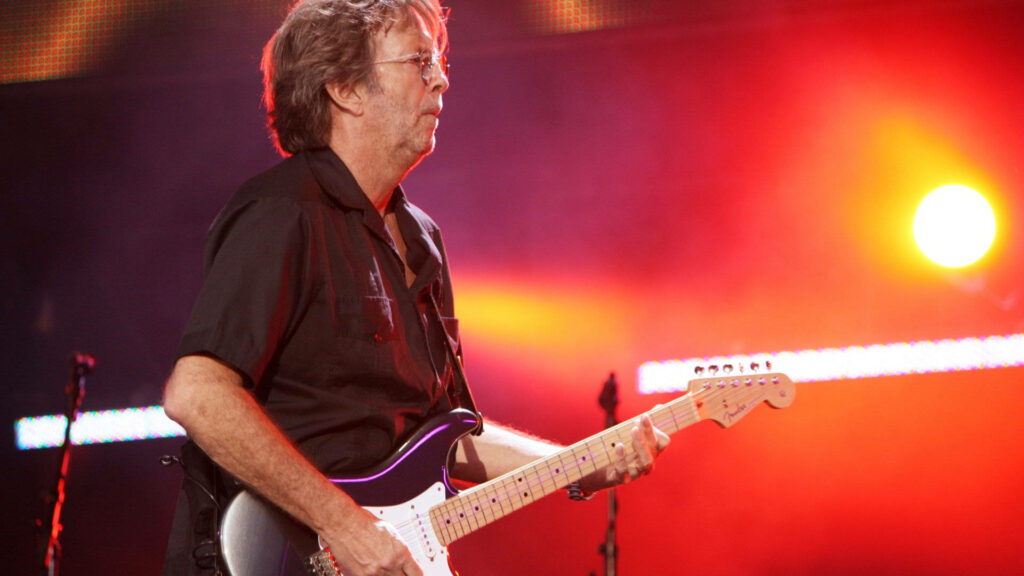 Eric Clapton passionately plays guitar on stage with vibrant orange light background wallpaper