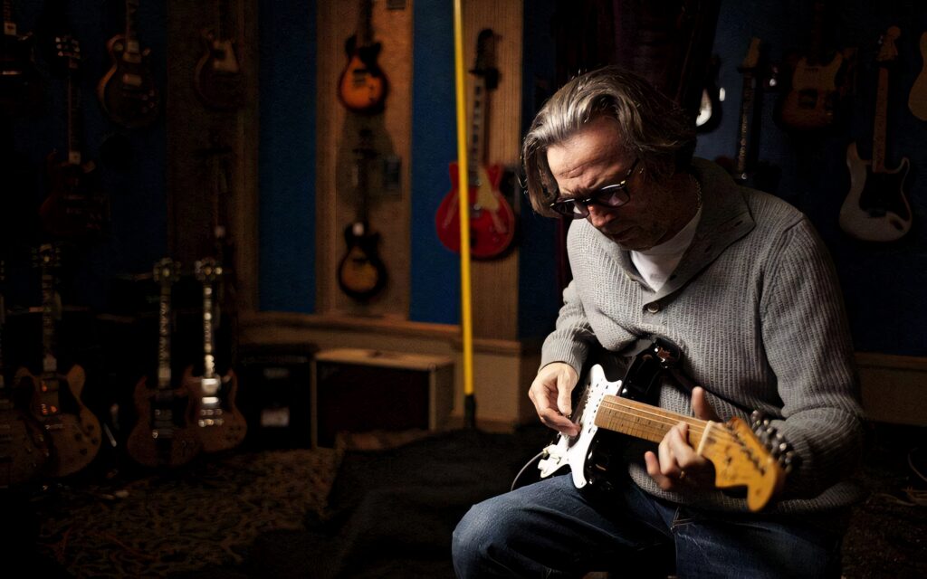 Eric Clapton playing electric guitar in a room filled with classic guitars on walls wallpaper