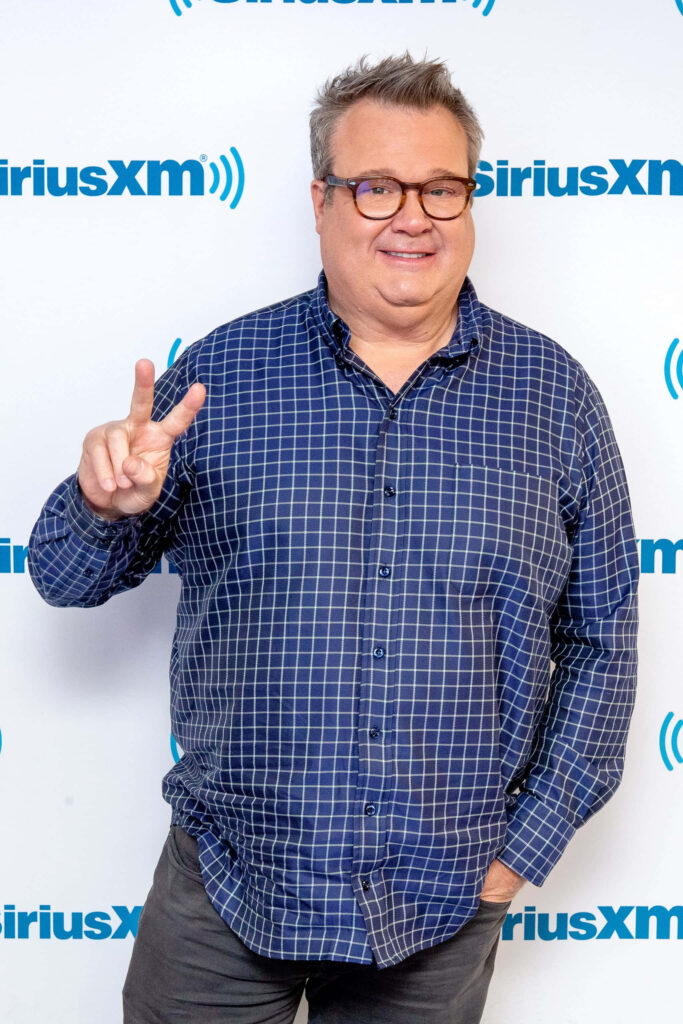 Eric Stonestreet smiling and flashing peace sign in blue checkered shirt at SiriusXM media event Wallpaper