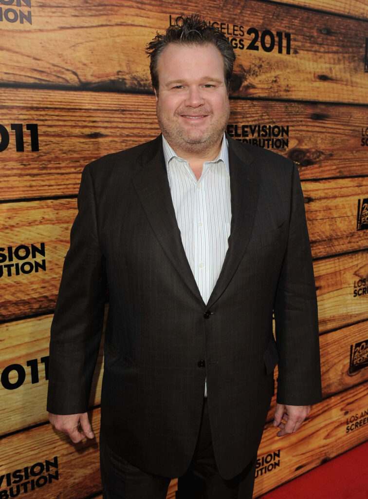 Eric Stonestreet in dark suit on red carpet with warm wooden backdrop and bold text wallpaper