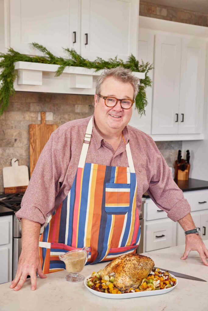 Eric Stonestreet in colorful apron presenting roasted turkey with vegetables in modern kitchen Wallpaper