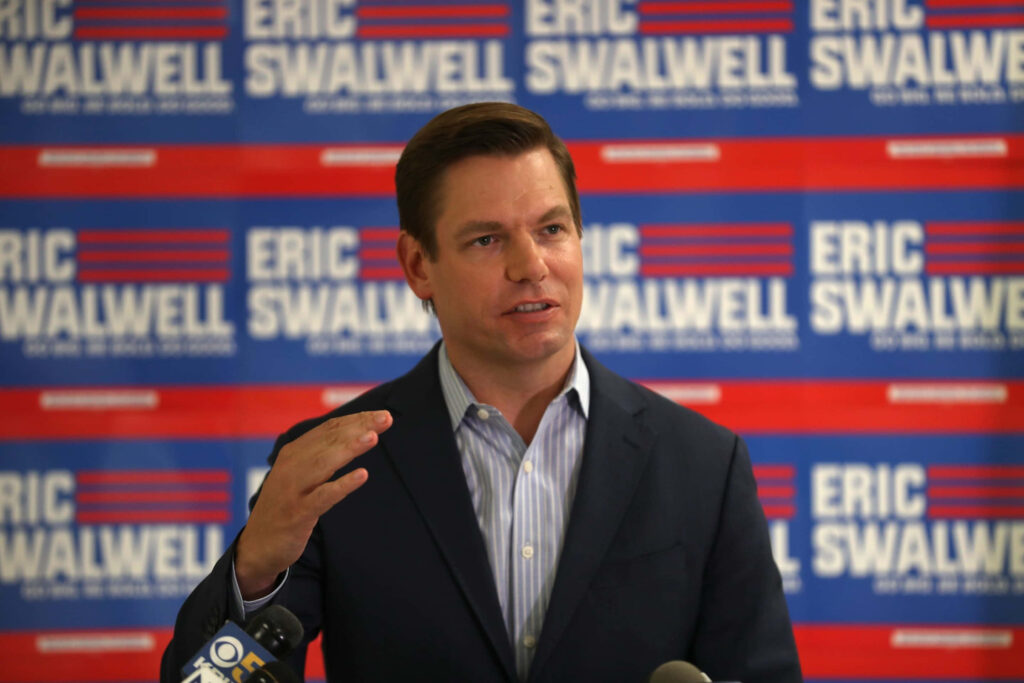 Eric Swalwell speaking confidently with campaign backdrop in red white and blue stripes wallpaper
