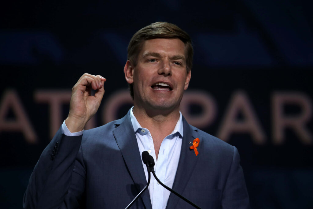 Eric Swalwell speaking on stage in dark suit with orange ribbon dramatic background wallpaper