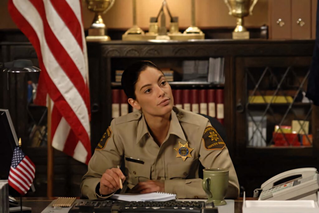 Erica Cerra as sheriff seated at desk with American flag law enforcement backdrop warm tone Wallpaper