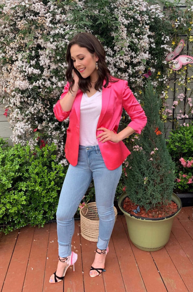 Erin Krakow in pink blazer and blue jeans on wooden deck with spring garden and white flowers Wallpaper