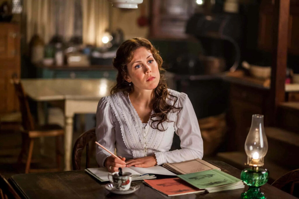Erin Krakow in vintage blouse seated at rustic wooden table with books and warm lighting Wallpaper