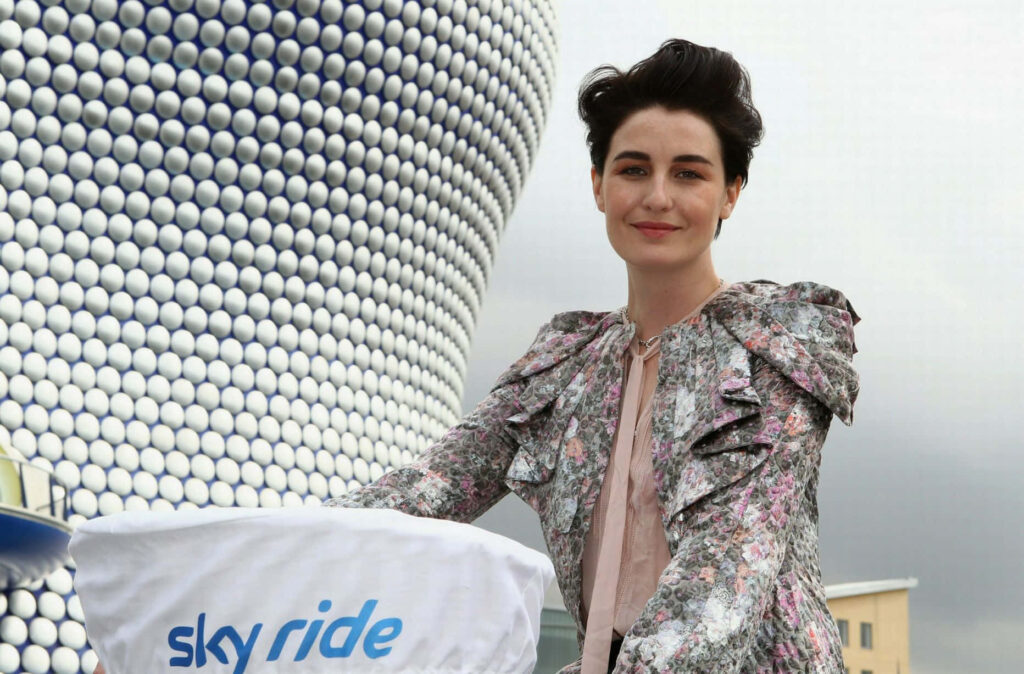 Erin OConnor in floral ruffled blouse stands before modern geometric circular architecture in daylight Wallpaper