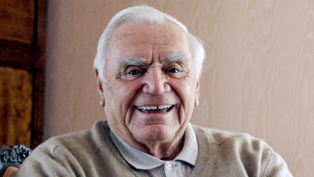 Ernest Borgnine smiling warmly in softly lit indoor setting with neutral background wallpaper
