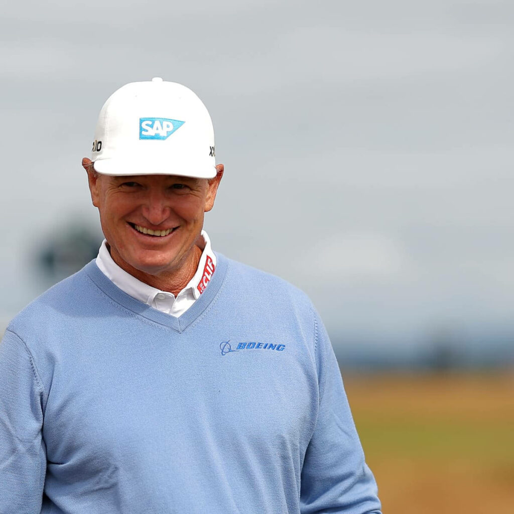 Ernie Els smiling in blue sweater and white cap with soft outdoor golf course background wallpaper