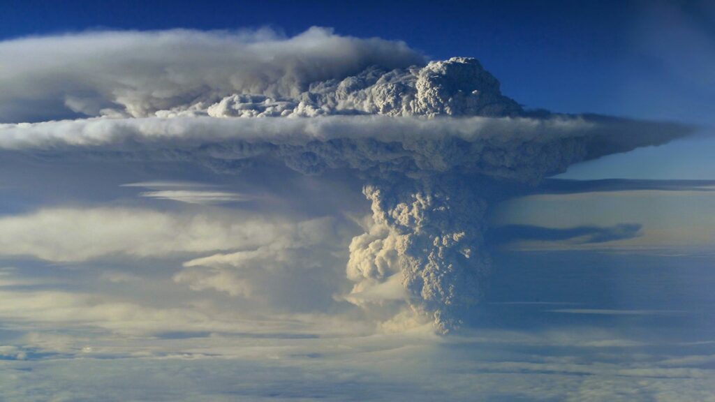 Volcano eruption with towering ash plume piercing clouds against bright blue sky wallpaper background