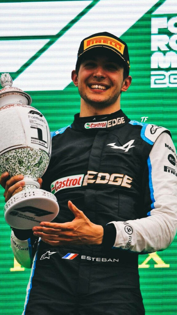 Esteban Ocon celebrating Formula 1 victory on podium with trophy and Pirelli cap against green backdrop Wallpaper
