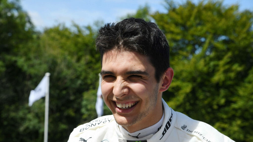 Esteban Ocon smiling outdoors in racing suit with sponsor logos against green natural backdrop Wallpaper