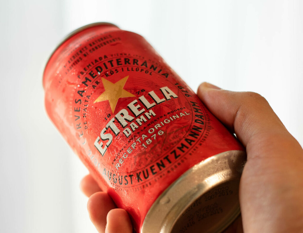 Hand holding vibrant red Estrella Damm can with bold star logo and crisp condensation on white background Wallpaper