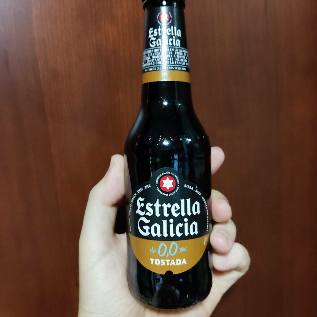Hand holding Estrella Galicia Dark 0 0 Tostada beer bottle with brown gold label on wood background Wallpaper