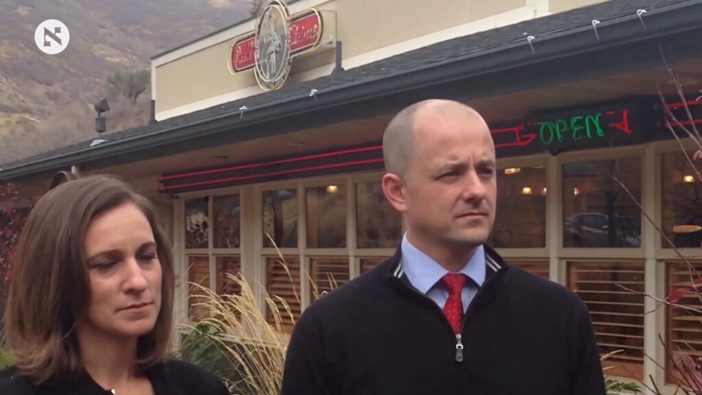 Evan McMullin and woman in formal attire outside rustic restaurant with mountain wallpaper background