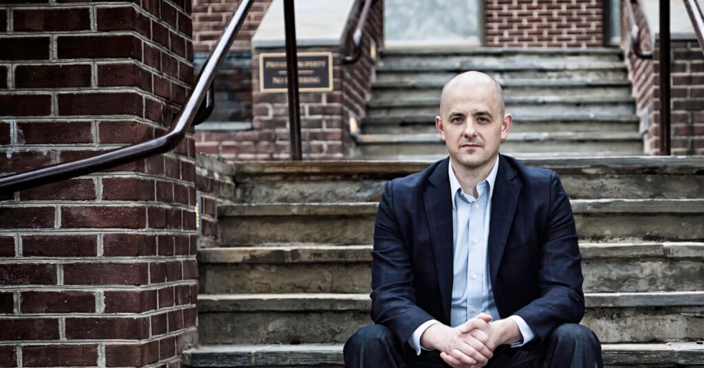 Evan McMullin seated on stone steps with red brick background showing leadership and focus Wallpaper