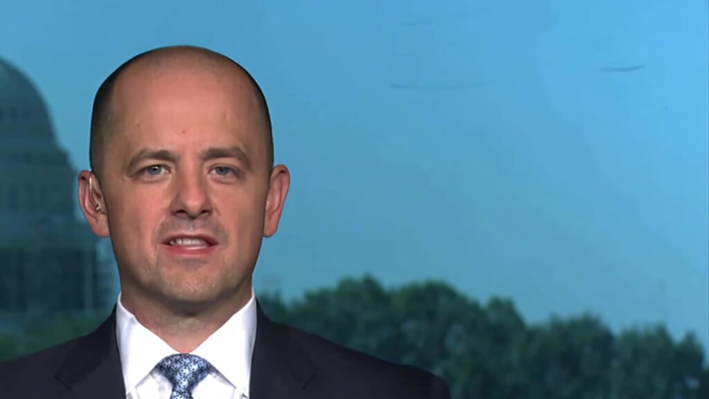 Evan McMullin in formal suit with blurred government building and soft blue political wallpaper background