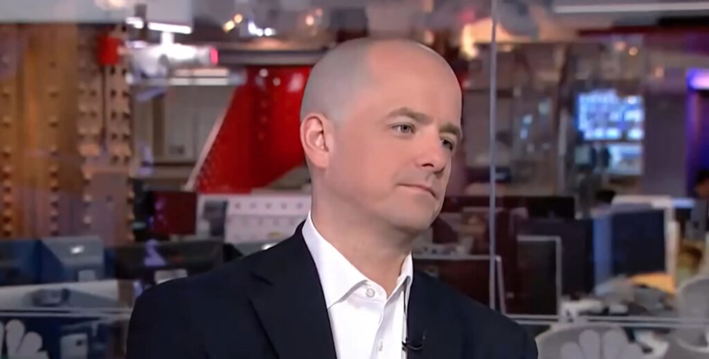Evan McMullin in formal suit in modern newsroom with glass panels and muted lighting wallpaper