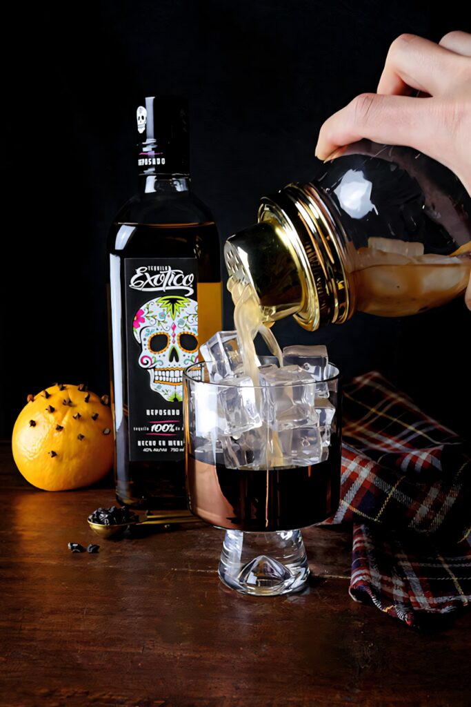 Hand pouring Exotico Tequila orange juice mix over ice with bottle and spiced orange on wood table Wallpaper