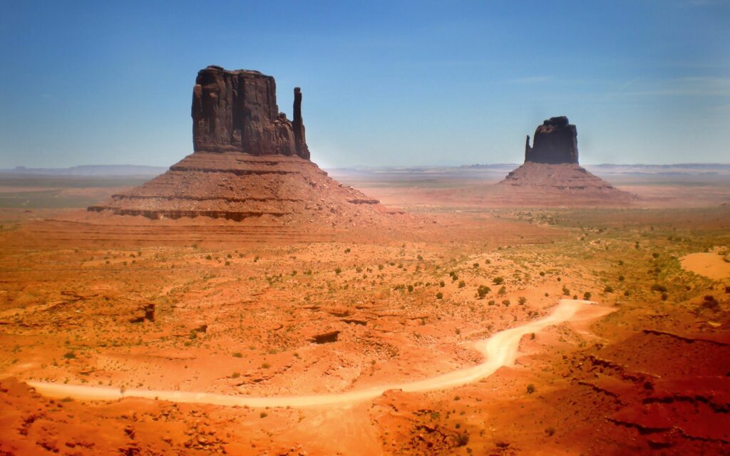 Expansive desert landscape with towering red rock canyons and sparse green vegetation under blue sky Wallpaper
