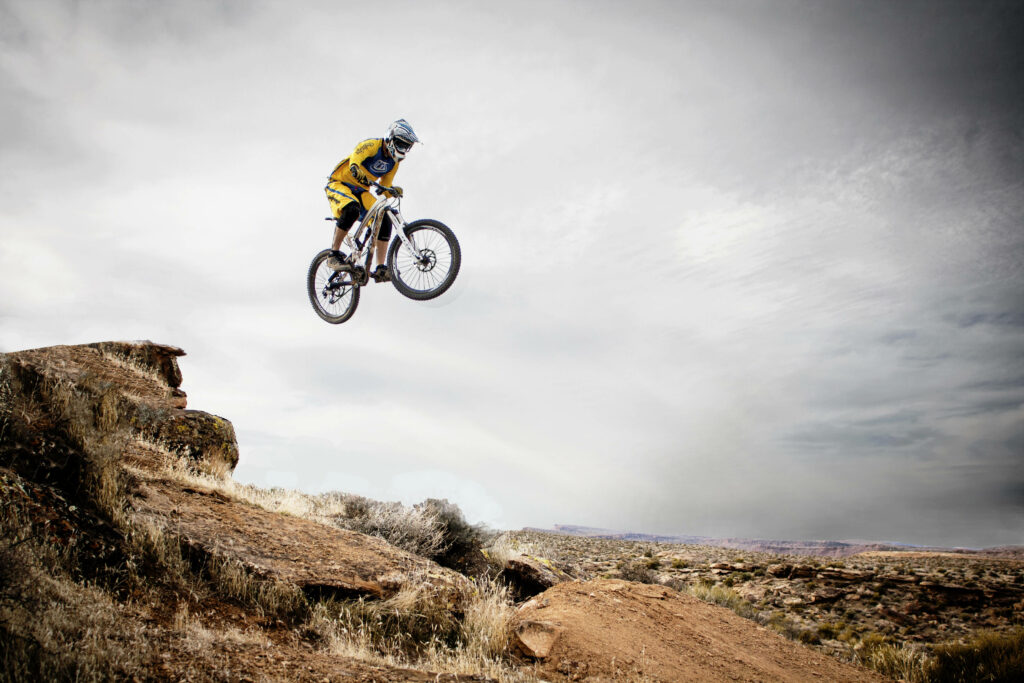Cyclist in yellow gear jumping off rugged rock ledge extreme mountain biking adventure outdoors Wallpaper