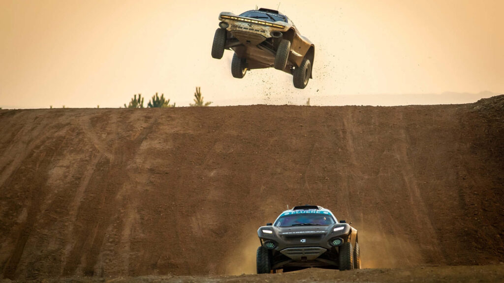 Two off-road vehicles race in a desert one airborne over dusty ramp extreme desert race wallpaper