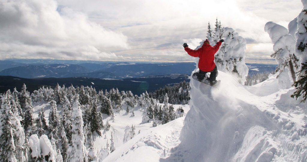 Extreme snowboarding jump from snowy mountain peak with rider in red jacket and pine trees Wallpaper
