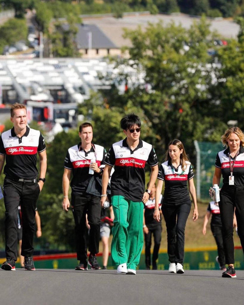 F1 driver Zhou Guanyu in bright green pants with Alfa Romeo team at racetrack paddock daylight Wallpaper