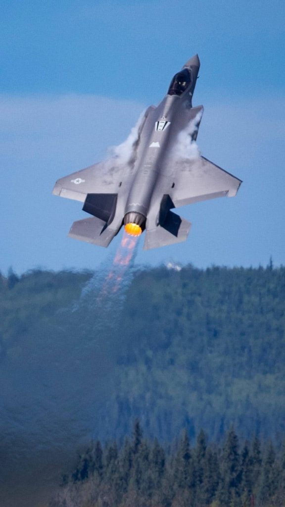 F-35 Lightning II fighter jet ascending with glowing afterburner over forest clear sky wallpaper