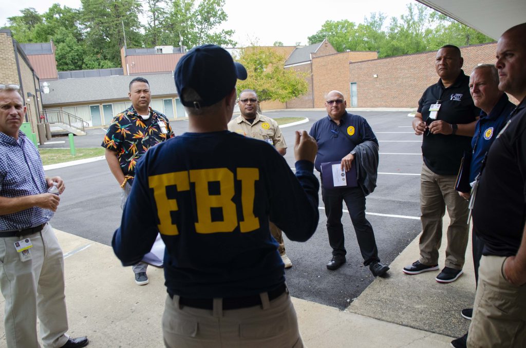 FBI agent in navy shirt briefing diverse law enforcement officers outdoors in daylight parking lot Wallpaper