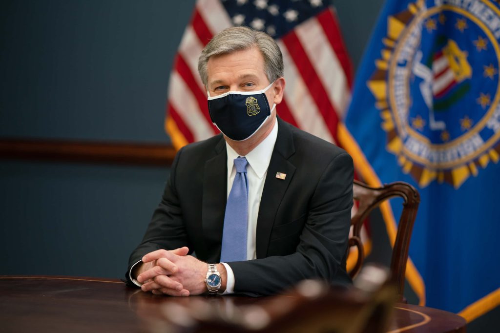 FBI Director Christopher Wray in dark suit with mask and flags in official government office setting Wallpaper