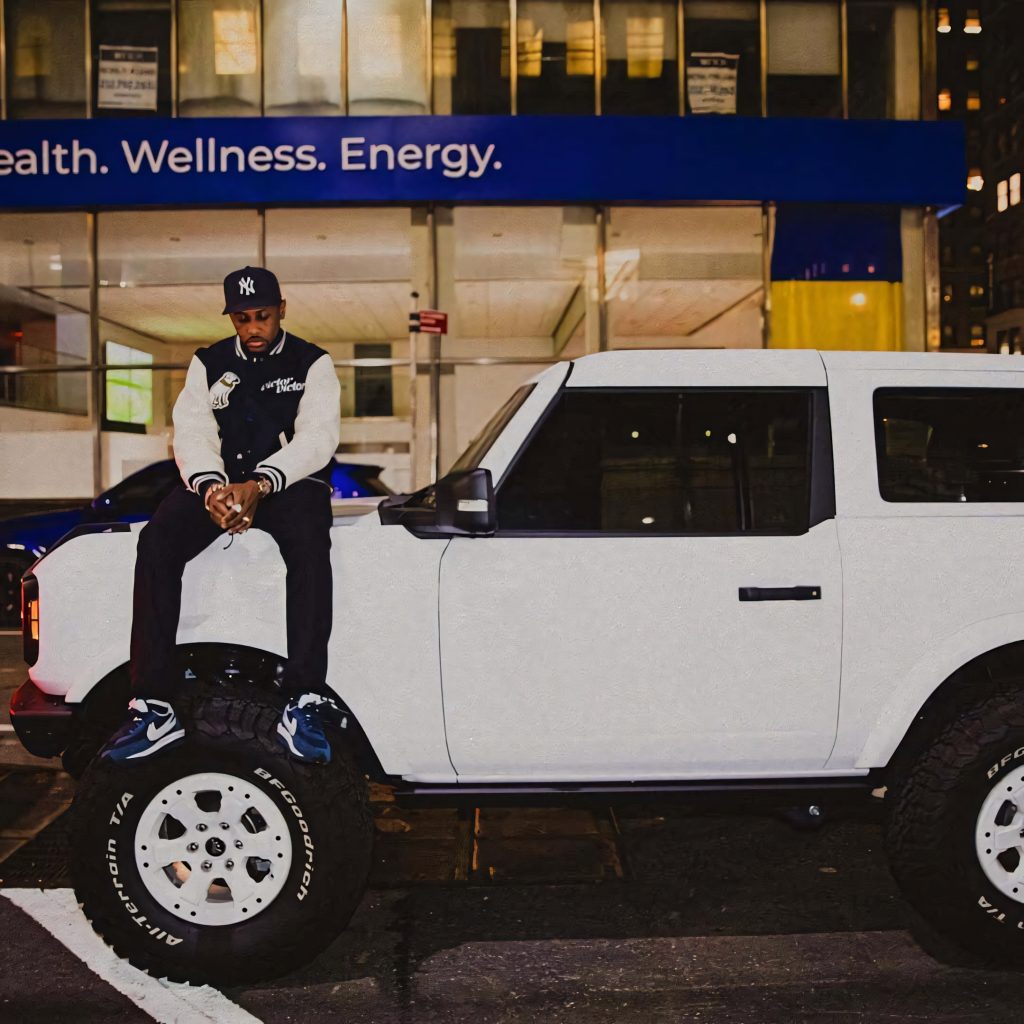 Fabolous rapper in varsity jacket sitting on rugged white SUV with off-road tires under city lights Wallpaper