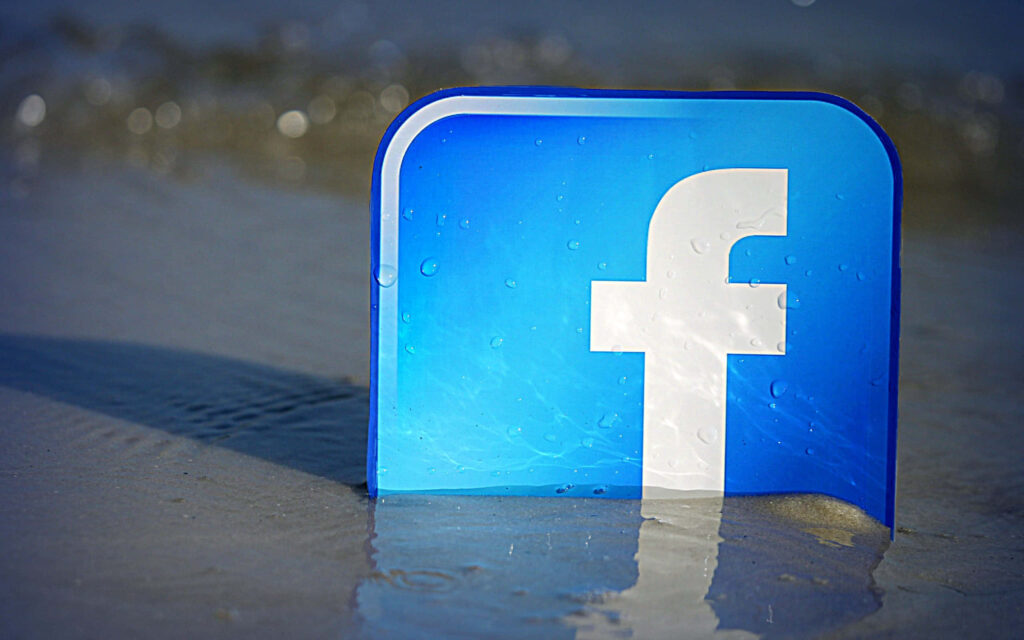 Bold blue Facebook logo on sandy shore with gentle waves blending social media and nature Wallpaper