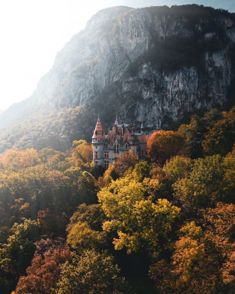 Charming castle surrounded by vibrant autumn trees rugged rocks and towering mountain backdrop Wallpaper