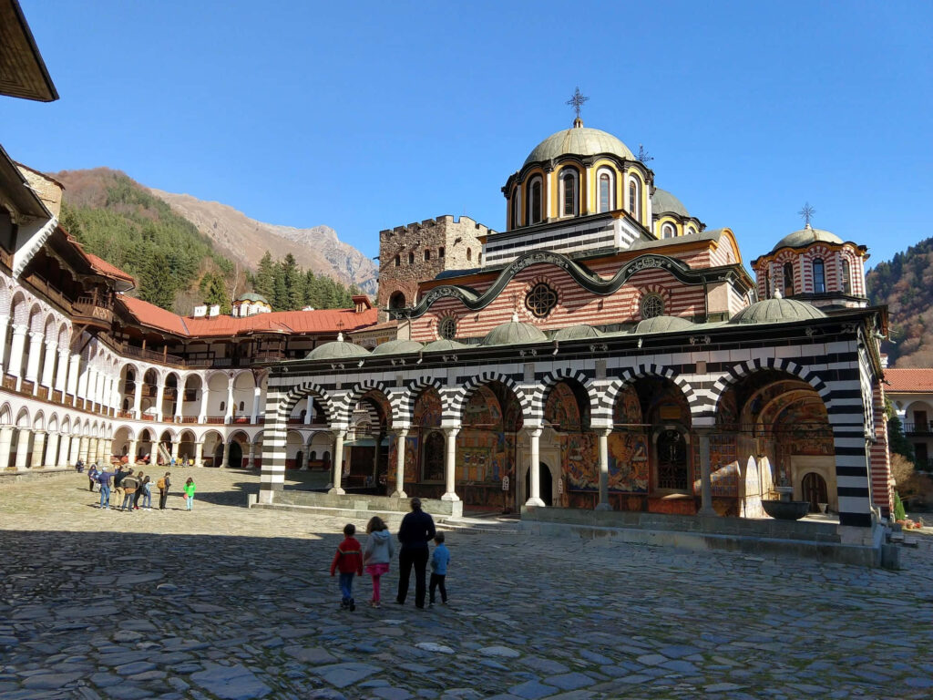 Family visiting historic Rila Monastery with mountain views and Bulgarian architecture under blue sky Wallpaper