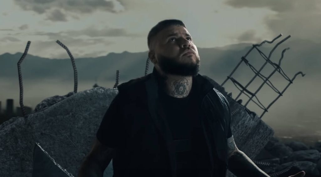 Farruko in black stands reflective among concrete rubble under a moody gloomy sky wallpaper background