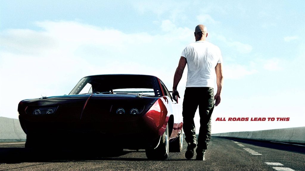 Vin Diesel walking on sunlit highway beside red classic muscle car Fast and Furious poster wallpaper