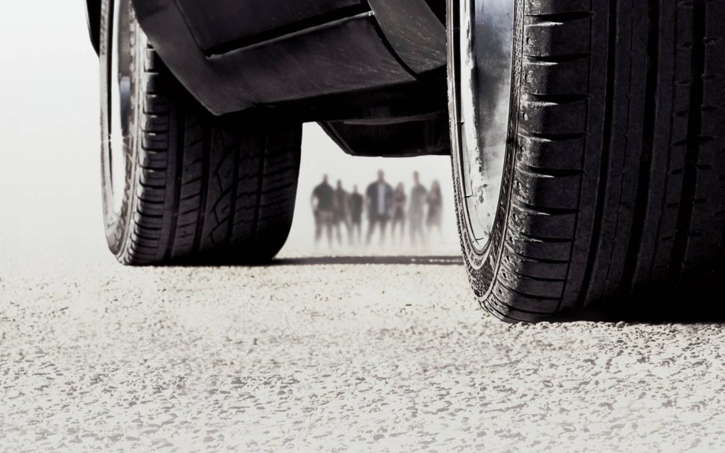 Low-angle close-up of muscle car tires gripping asphalt with Fast and Furious cast silhouettes background Wallpaper