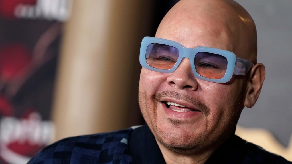 Fat Joe portrait smiling in oversized light blue sunglasses and dark textured shirt wallpaper background