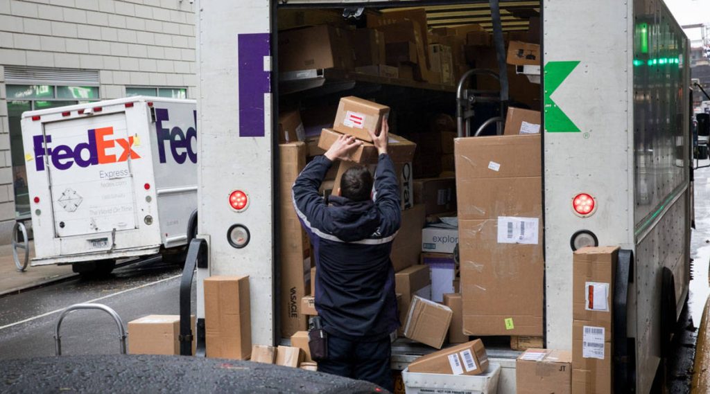 FedEx driver unloading packages from truck on busy street with second FedEx vehicle nearby Wallpaper