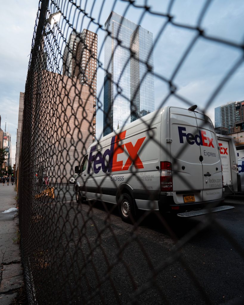 White FedEx Express delivery van parked behind chain-link fence with city skyscrapers background Wallpaper