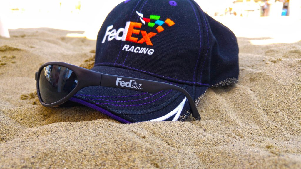 FedEx Racing black cap and sunglasses on sunny sandy beach vibrant purple sporty fan gear wallpaper background