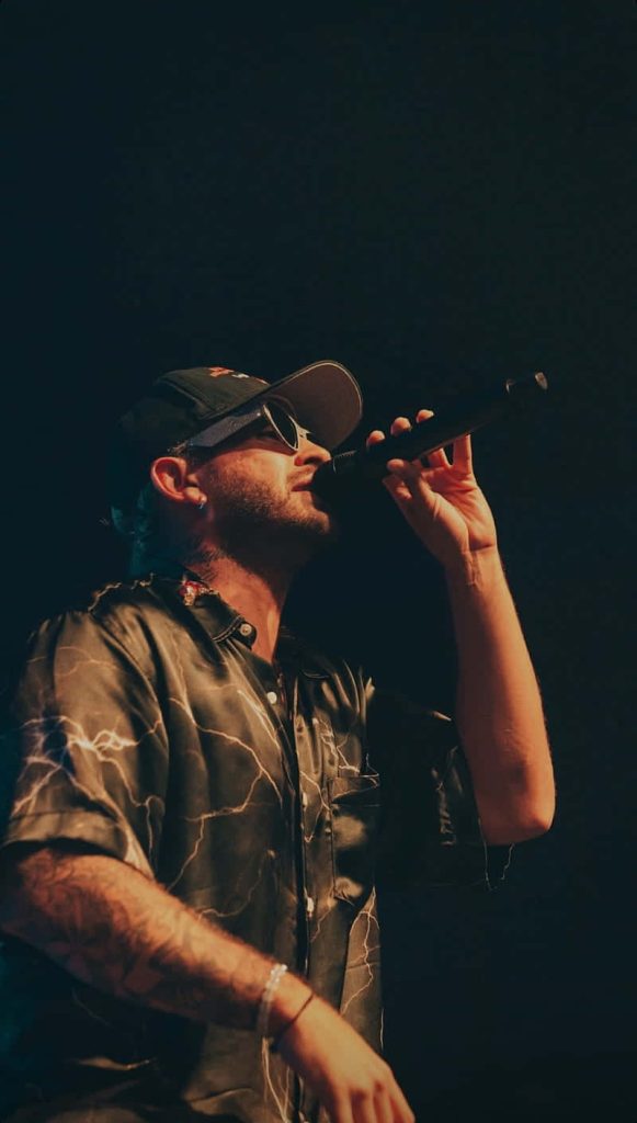 Colombian singer Feid live concert close-up with signature cap sunglasses and lightning shirt Wallpaper