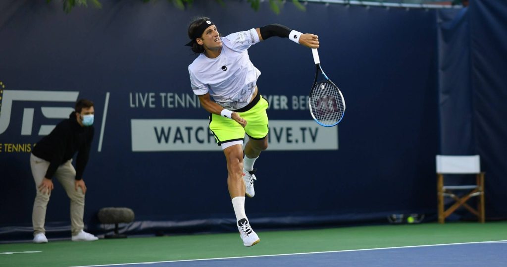 Professional tennis player Feliciano López mid-serve in white shirt neon shorts on blue court wallpaper