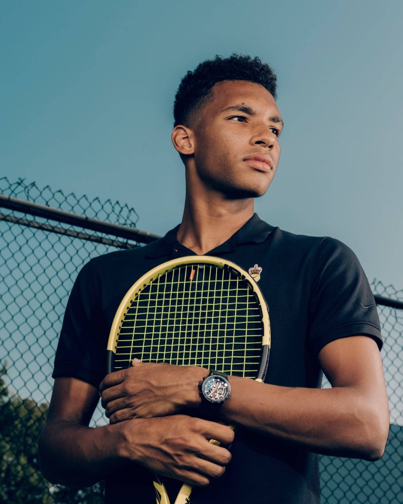Felix Auger-Aliassime tennis portrait black polo skeleton watch holding yellow racket fence background Wallpaper
